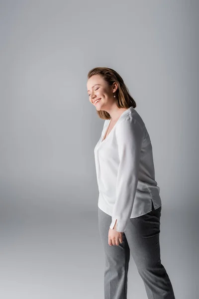 Sorridente, elegante ragazza sovrappeso in camicetta bianca in posa con gli occhi chiusi sul grigio — Foto stock
