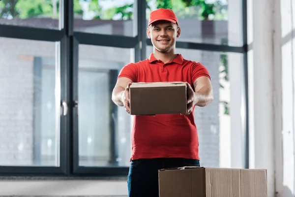 Happy delivery man in cap holding carton box and looking at camera — Stock Photo