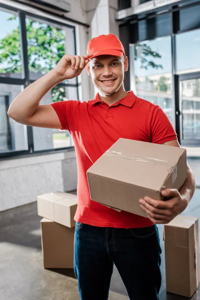 Glücklicher Zusteller berührt Mütze, während er Karton hält — Stockfoto