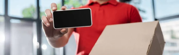 Panoramic crop of delivery man holding smartphone with blank screen and carton box — Stock Photo