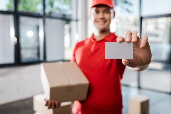 Foyer sélectif de l'homme de livraison heureux dans le chapeau tenant la carte blanche et la boîte de carton — Photo de stock