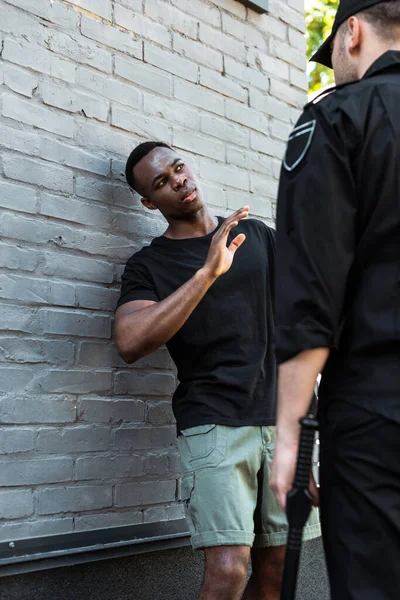 Enfoque selectivo del hombre afroamericano asustado con la mano levantada mirando al policía con bastón, concepto de racismo - foto de stock