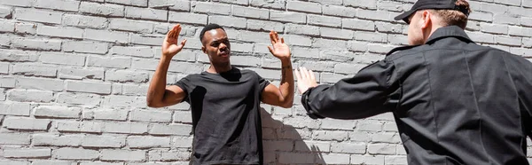 Panoramic orientation of policeman in cap detaining african american man with raised hands, racism concept — Stock Photo