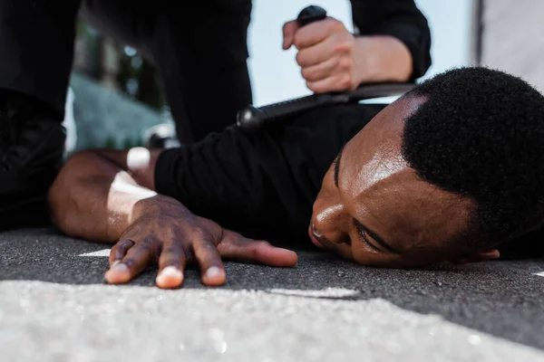 Enfoque selectivo del hombre afroamericano detenido acostado en el suelo cerca de la policía con bastón, concepto de racismo - foto de stock