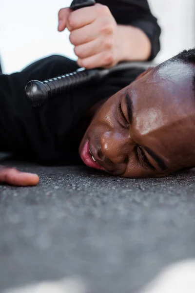 Enfoque selectivo del hombre afroamericano detenido acostado en el suelo cerca de la policía con porra, concepto de racismo - foto de stock