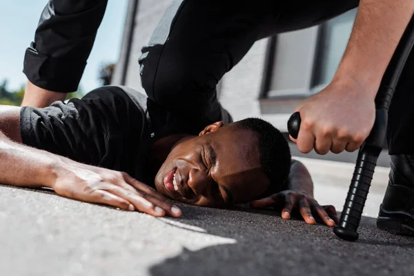 Enfoque selectivo de hombre afroamericano detenido y herido acostado en el suelo cerca de policía con truncheon, concepto de racismo - foto de stock