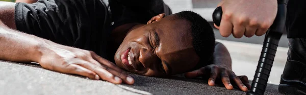 Horizontal image of detained and injured african american man lying on ground near policeman with truncheon, racism concept — Stock Photo