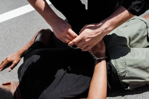 Visão cortada de policial segurando algemas enquanto detendo homem afro-americano na rua, conceito de racismo — Fotografia de Stock