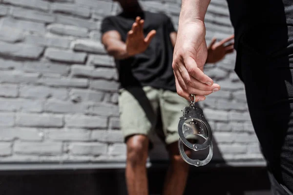 Cropped view of police officer holding handcuffs while detaining african american man gesturing near brick wall, racism concept — Stock Photo