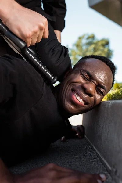 Selective focus of detained african american man suffering from pain while lying on ground near policeman with baton, racism concept — Stock Photo