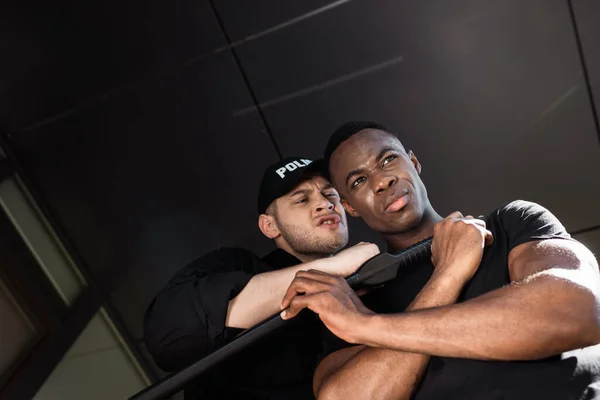 Low angle view of angry police officer holding truncheon near african american man, racism concept — Stock Photo