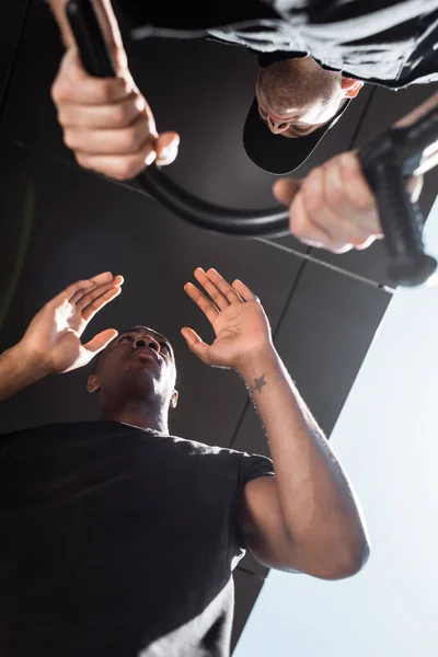 Vista inferior del policía sosteniendo porra cerca del hombre afroamericano tatuado, concepto de racismo - foto de stock