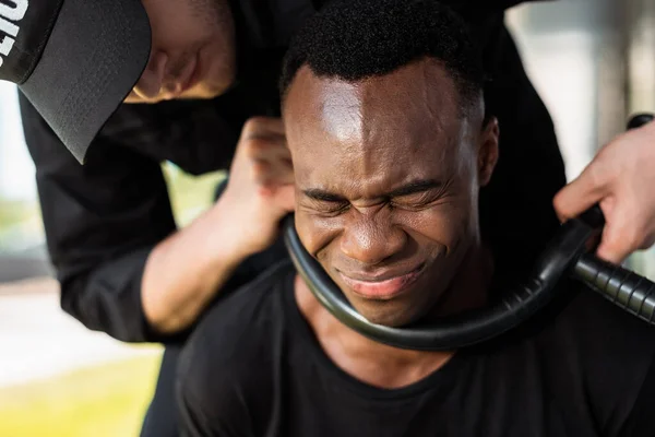 Policeman holding police baton near throat of scared african american man with closed eyes, racism concept — Stock Photo