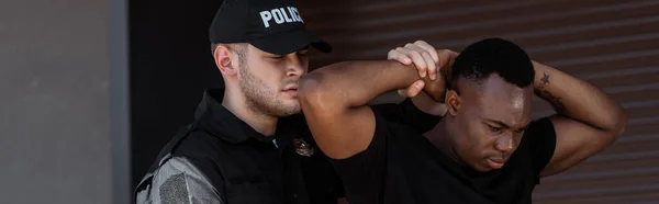 Prise de vue panoramique d'un policier en casquette avec un lettrage de police détenant un homme afro-américain — Photo de stock