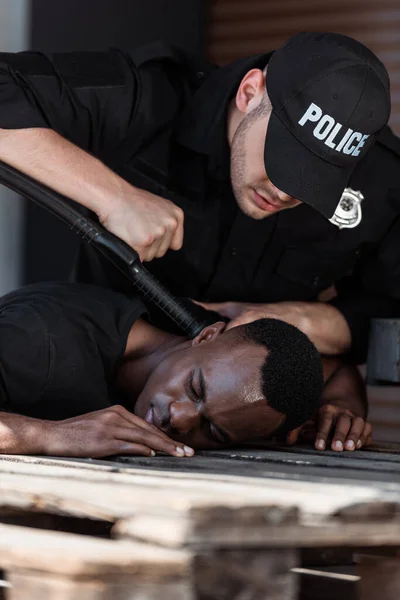 Foco seletivo de policial em cap com letras policiais segurando truncheon enquanto detendo homem afro-americano — Fotografia de Stock