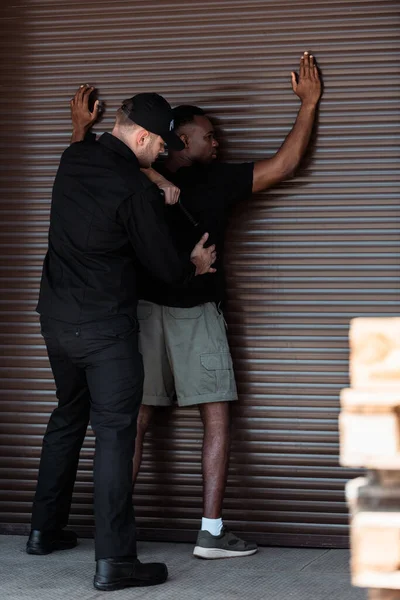 Selective focus of police officer in uniform and cap holding truncheon while detaining african american man — Stock Photo