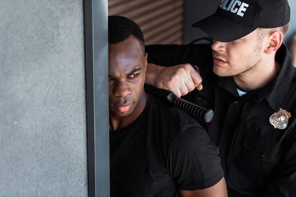 Enfoque selectivo del oficial de policía en uniforme y gorra que sostiene la porra mientras arresta al hombre afroamericano - foto de stock