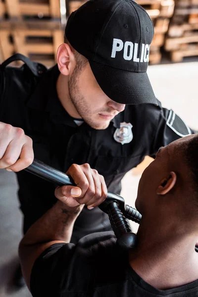 Angry police officer in cap with police lettering holding truncheon while arresting african american man — Stock Photo