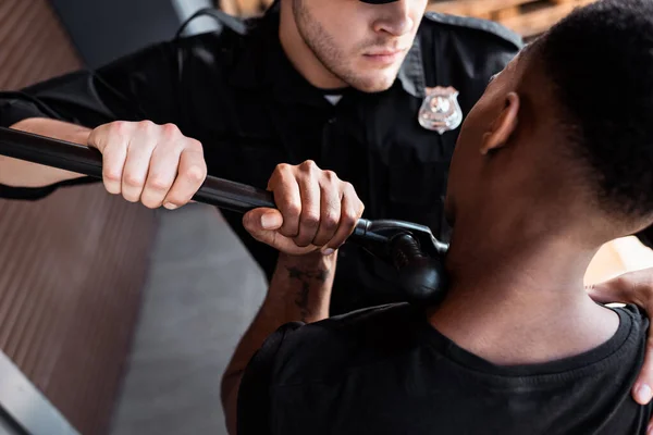 Vista cortada de policial irritado segurando truncheon enquanto prendia homem afro-americano — Fotografia de Stock