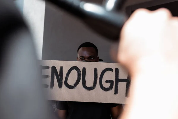 Foyer sélectif de l'homme afro-américain avec écharpe sur le visage tenant la plaque avec assez de lettrage près de policier, concept de racisme — Photo de stock