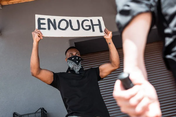 Selective focus of african american man with scarf on face holding placard with enough lettering near policeman with truncheon, racism concept — Stock Photo