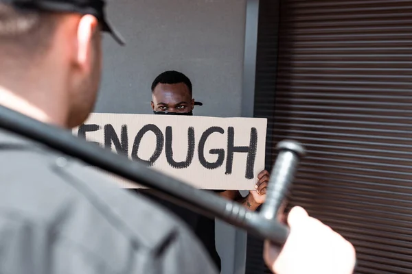 Selective focus of african american man holding placard with enough lettering near police officer with truncheon, racism concept — Stock Photo