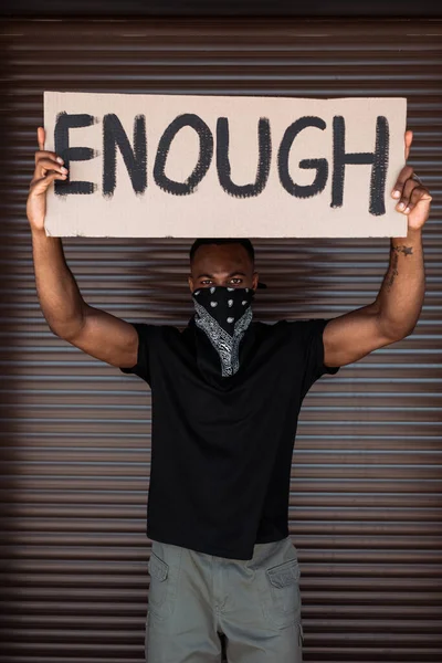 Hombre afroamericano con bufanda en la cara sosteniendo pancarta con suficientes letras en la calle, concepto de racismo - foto de stock