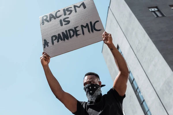 Low angle view of african american man with scarf on face holding placard with racism is a pandemic lettering near building — Stock Photo