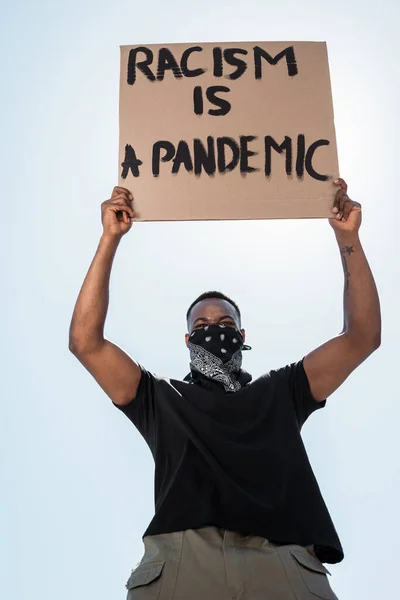 Low angle view of tattooed african american man with scarf on face holding placard with racism is a pandemic lettering against blue sky — Stock Photo