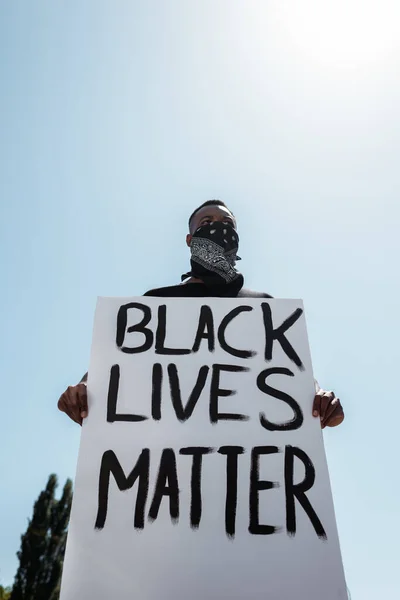 Vue à angle bas de l'homme afro-américain avec écharpe sur le visage tenant la plaque avec des vies noires matière lettrage contre ciel bleu — Photo de stock