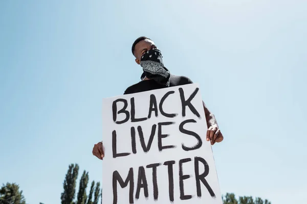 Vista de ángulo bajo del hombre afroamericano con bufanda en la cara sosteniendo pancarta con letras de materia de vidas negras contra el cielo - foto de stock