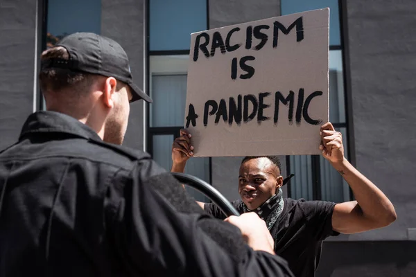 Enfoque selectivo del hombre afroamericano con pancarta con racismo es una letra pandémica cerca de policía con porra en la calle - foto de stock