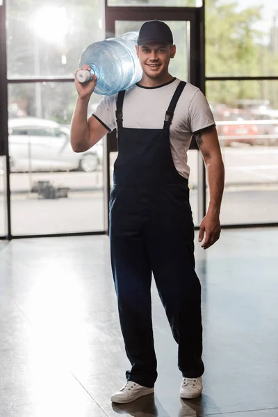 Heureux livreur homme en capuchon et uniforme tenant bouteille d'eau — Photo de stock