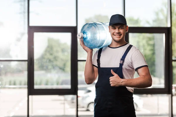 Heureux livreur homme en uniforme tenant bouteille d'eau et montrant pouce vers le haut — Photo de stock