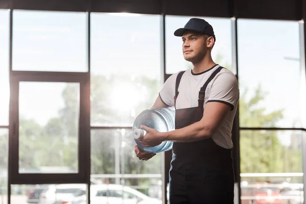 Handsome delivery man in uniform and cap holding bottled water — Stock Photo