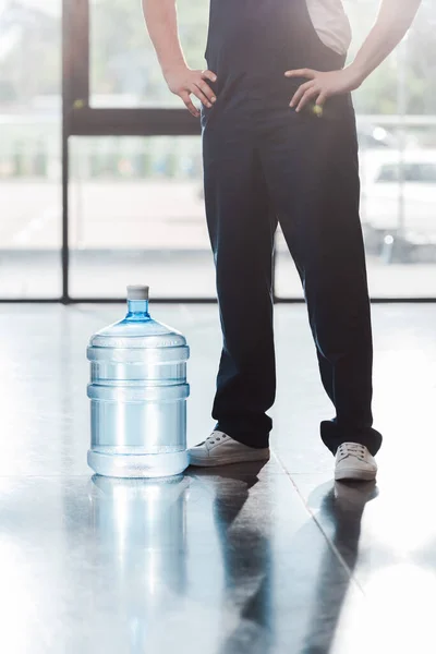 Vista recortada del repartidor en uniforme de pie con las manos en las caderas cerca de agua embotellada — Stock Photo