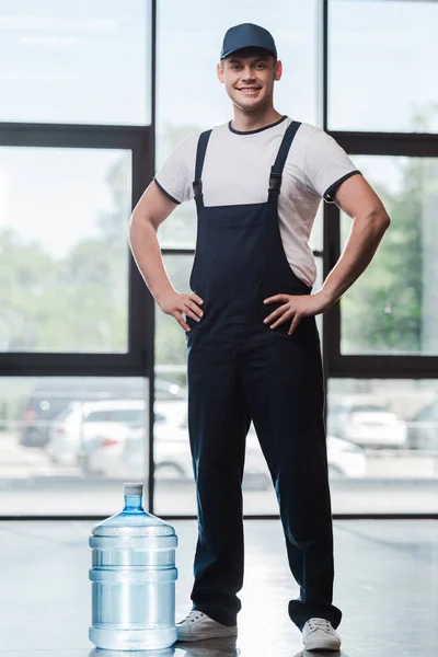 Веселий чоловік доставки в уніформі, що стоїть руками на стегнах біля пляшкової води — стокове фото