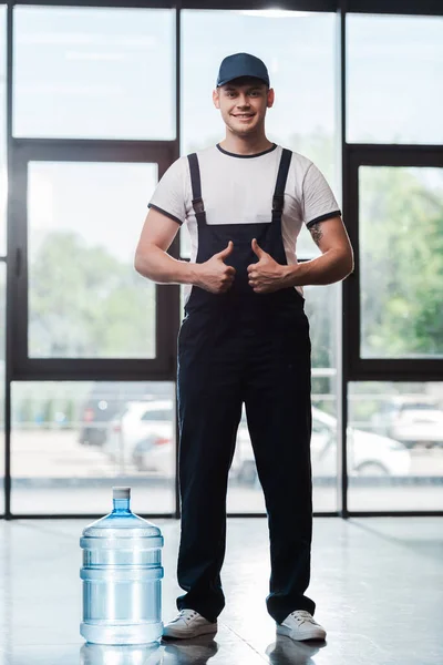 Alegre repartidor en uniforme mostrando pulgares hacia arriba cerca de galón de agua embotellada - foto de stock