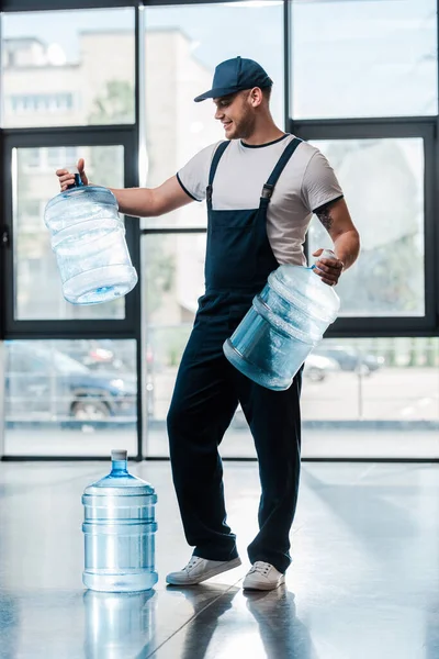 Alegre repartidor en uniforme mirando botellas vacías cerca de galón de agua - foto de stock