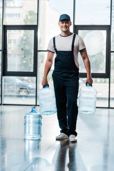 Веселий чоловік доставки в уніформі тримає порожні пляшки біля галон води — стокове фото