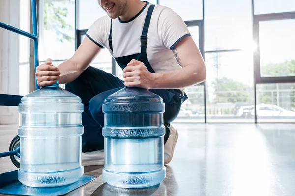 Cropped view of happy delivery man holding blue bottles with water near hand truck — Stock Photo