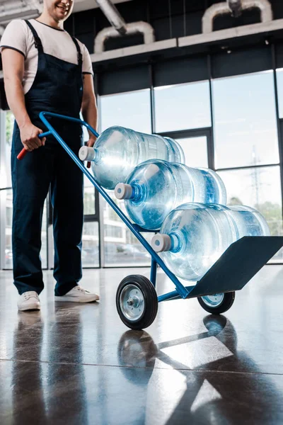 Cropped view of cheerful delivery man in uniform holding hand truck with bottled water — Stock Photo