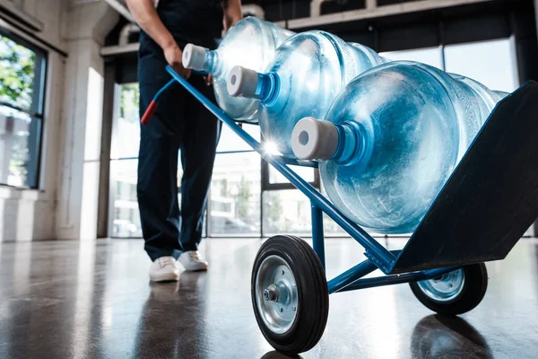 Cropped view of delivery man in uniform holding hand truck with purified water in bottles — Stock Photo