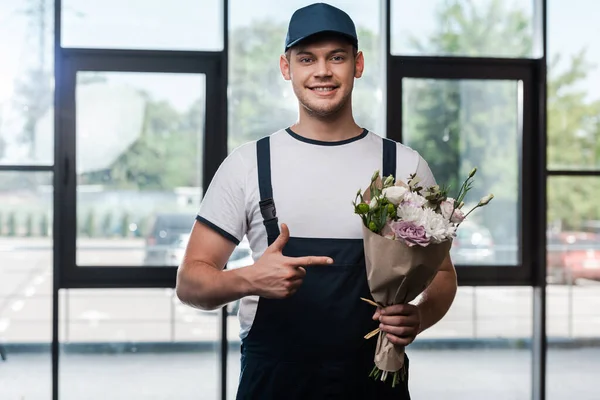 Heureux livreur homme en uniforme et chapeau pointant du doigt au bouquet avec des fleurs — Photo de stock