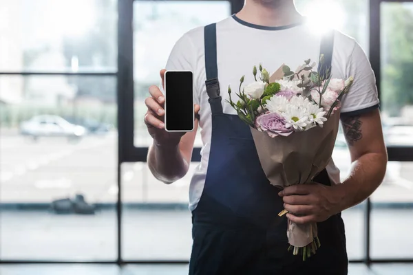 Cropped view of delivery man holding bouquet of blooming flowers and smartphone with blank screen — Stock Photo