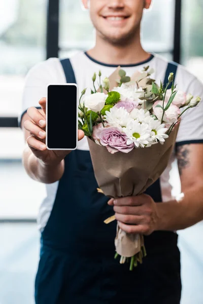 Cropped view of happy delivery man holding bouquet of blooming flowers and smartphone with blank screen — Stock Photo