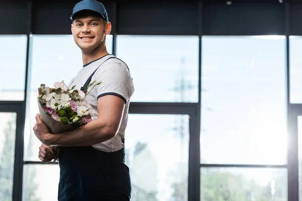 Felice uomo di consegna in cappello e uniforme che tiene bouquet di fiori in fiore — Foto stock