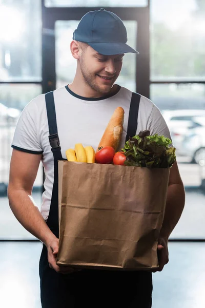 Happy delivery man in cap looking at paper bag with groceries — Stock Photo