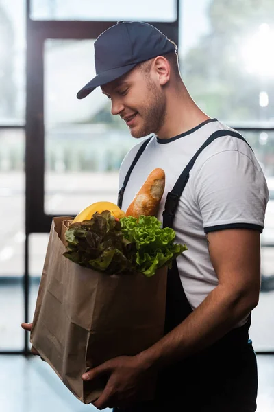 Alegre repartidor hombre en gorra mirando bolsa de papel con comestibles - foto de stock
