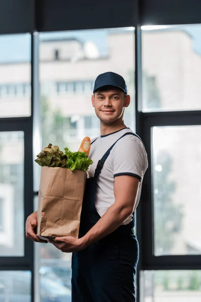 Alegre repartidor hombre en gorra sosteniendo bolsa de papel con comestibles - foto de stock